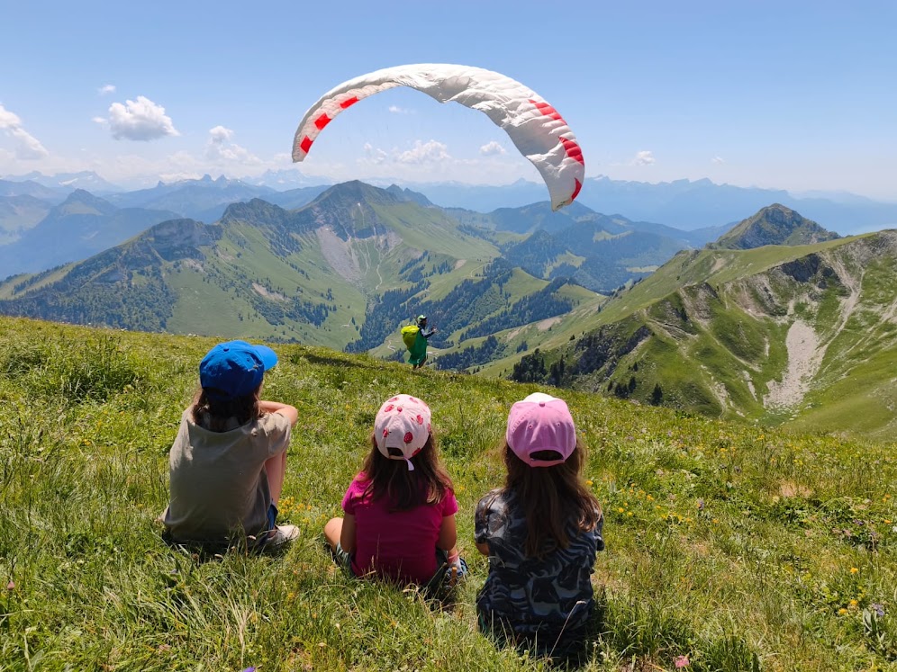 Trois enfants, de dos, regardent un décollage face aux Préalpes.