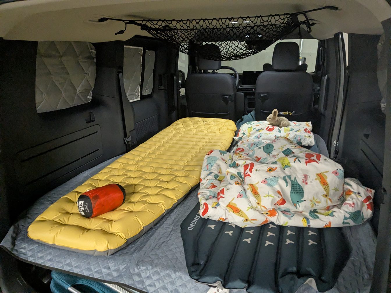 Le lit fait dans le van : deux matelas gonflables, une couette à motifs d'oiseaux, une peluche lapin, et des rideaux isolants aux fenêtres.