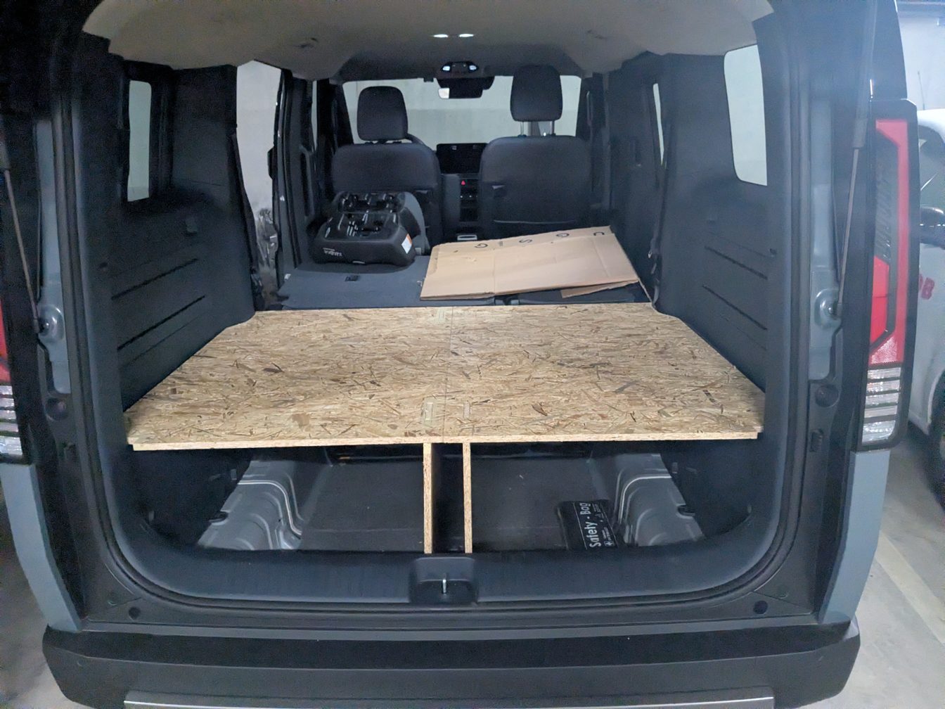 Le coffre du van ouvert, sièges arrière rabattus : un plateau en panneau OSB posé sur deux pieds en bois, avec un espace de rangement dessous.